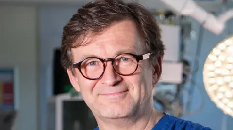 A man with brown hair and round brown glasses wearing blue scrubs is in an operating theatre.