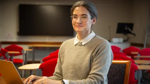 Dr Robyn Jackowich has glasses and grey hair tied back behind her ears. She is sitting at a laptop, in an office. Smiling at the camera. A TV is mounted on the wall behind her and there a several red chairs in the background 