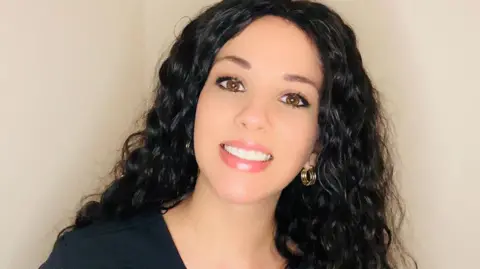 Anastasia Cameron Anastasia, with long, curly black hair, is smiling at the camera and standing against a blank wall