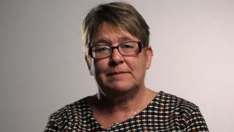 Roxy looks into the camera with a serious expression. She has short blonde hair, wears dark framed glasses and a black and white patterned top with a round neck and a small silver chain around her neck