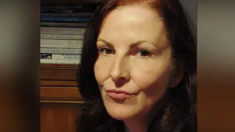 Debbie Foster A woman with dark red hair, wearing a black cardigan over a read and black striped top looks into camera as she sits in a dimly lit room.