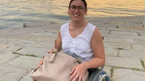 Sofia Brizio A woman in her twenties sits in a wheelchair with a large bag on her lap next to water, she smiles and looks towards the camera