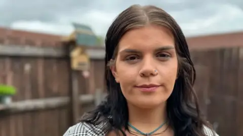 BBC Sophie Henson looks into the camera. She has brown hair and dark eyes, is in her garden and wears a blue necklace. 