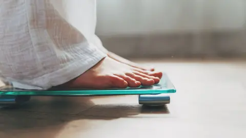 Getty Images Person weighing themselves on scales. A set of scales can be seen on the floor of a bathroom. The feet of a person is in view as they step on the scales.