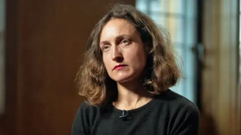 Headshot of Cécile Simmons. She has shoulder-length brown wavy hair and a black crew-neck jumper. She is pictured in a wood-panelled room