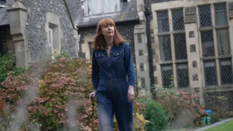 Elise Burns walking in a garden with a walking stick. She is wearing a denim outfit and has light brown hair.