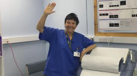 HANDOUT Jane Graham, wearing dark blue hospital scrubs and a lanyard, waves in a hospital consultation room