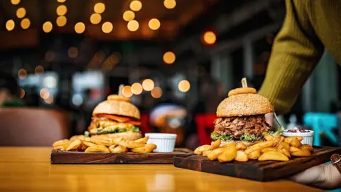 Getty Images Two wooden boards, containing large burgers and chunky chips and sauces, are being delivered to a restaurant table in the evening, while lights are on in background