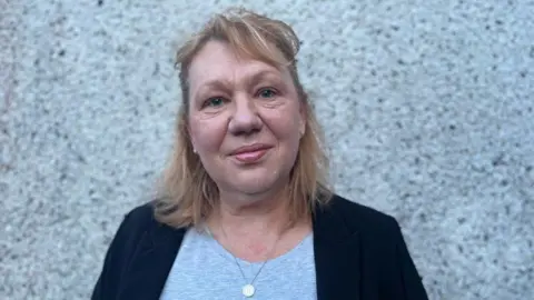 Helen Whyley smiling at the camera. She has short blonde hair and is wearing a black blazer and grey top. 