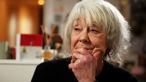 Wendy is pictured - a head and shoulders shot. She is 80, with tousled straight grey hair. Her chin is resting on her left hand, and she is wearing a gold and amber ring on her ring finger. She is looking off to her right. 