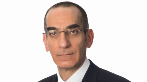 A man who is about 50, with short dark shaved hair, distinctive dark eyebrows and rimless glasses. He is looking straight at the camera and is not smiling. He is pictured - head and shoulders - against a plain white background. 