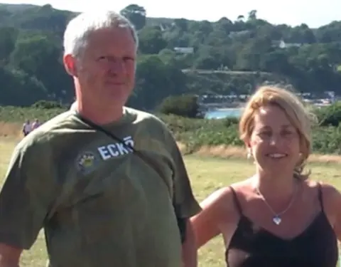 Julie Rhodes A man and a woman are pictured in the countryside by an expanse of water. It is a sunny summer's day and trees are in full leaf on the hillside in the distance. The man is wearing a green T-shirt, he has straight grey hair and is smiling at the camera. The woman is also smiling. She has straight blond hair, is wearing a necklace with silver heart, and is wearing a brown summery top with thin shoulder straps.