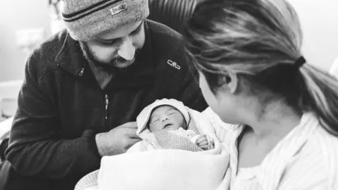 Family photo Black and white photo of Amarjit and Mandip holding their newborn baby Asees. She is wrapped in a blanket with a hat on. She is in Amarjit's arms and both parents are looking down at her. 