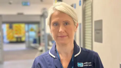 BBC A blonde haired women wears a navy nurse uniform, behind her is an ambulance 