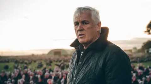 Paul Kelly A farmer with medium length white hair and a dark green quilted jacket stands in a portait. In the background but blurred out of focus are dozens of turkeys. They are among green grass and there's a type of domed green outhouse in the far distance.