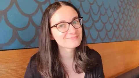 Nell A woman sat down on a bench that is against a wall. She is smiling to camera and is wearing glasses. Behind her is blue wallpaper. 