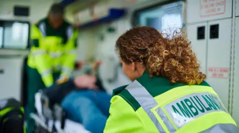 Getty Images Two paramedics load a stretcher bearing a patient into an ambulance - one, in the foreground, is wearing a yellow luminous jacket with the word Ambulance on the back of it.