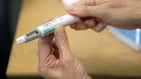 A pair of hands holding a Wegovy injection pen, which consists of a white plastic body with a dial and a window and a needle at the end. The label reads "Wegovy flextouch" with a dose of 0.25mg, and "injection semaglutide".