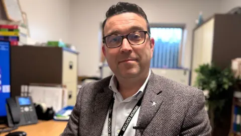 Dr Christopher Southwell has dark short hair and tortoise shell glasses on. He is wearing a grey tweed blazer and a pale blue shirt with a black lanyard that says "senior doctor". He is sitting at a desk which has a computer and phone on it.