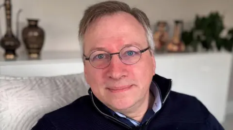 BBC A man with round glasses and short grey hair sits on a silver sofa, looking down the lens of the camera. He has his lips pursed and his eyes are a piercing blue colour. He is wearing a navy quarter zip jumper with a silver zip.