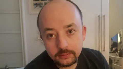 Roisin Wilshaw Brian Rooney has short dark hair and a goatee, he is wearing a black t-shirt and looking into the camera - he is sitting in a room with a cream wardrobe behind him 