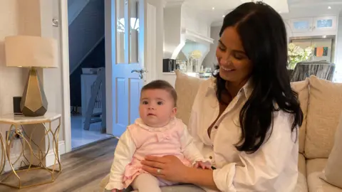 A dark-haired woman with a baby on her knee. The woman is smiling and wearing a white shirt. The baby girl is looking ahead and is wearing a pink pinafore with a cream top underneath and cream tights. The pair are sitting on a grey sofa with a gold table and lamp to one side of them and a pale coloured kitchen in the background.