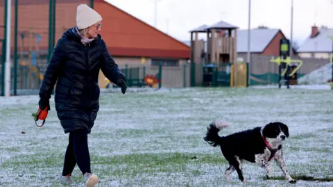 PA Media A woman wearing a hat and puffer jacket walks next to a dog. There's a dusting of snow on the ground