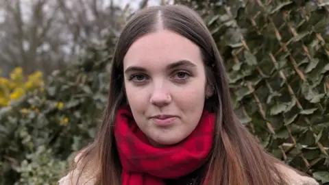 Mollie looks straight ahead, standing in front of a some greenery - she has long brown hair parted in the middle and wears a red scarf