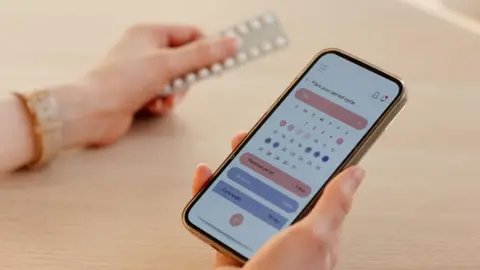 Getty Images A woman checks her calendar on her mobile phone while holding a packet of contraceptive pills in her other hand