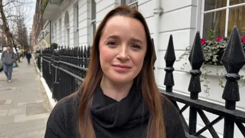 BBC 25-year-old Sophie stands outside an office on the pavement in front of black railings with a smile on her face.