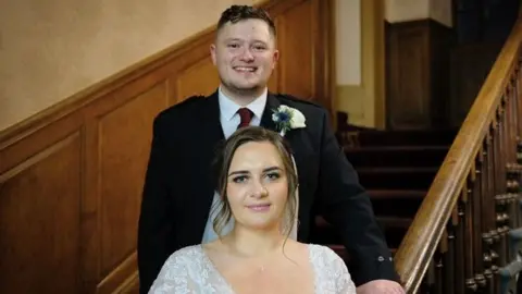 Weronika Somerville The couple pose on a stairway in the wedding venue which has wood-panelled walls and bannisters. Cameron stands behind his bride - he is wearing a kilt, jacket, burgundy tie and has a flower in his buttonhole while Weronika is in a lace gown and veil.