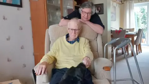 Diana Gibb Mick sits in an armchair stroking his black labrador dog with his walking aid beside him. His wife Diana stands behind him smiling.