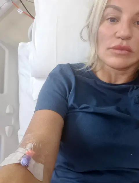 A selfie of Joanne in hospital, with a canula in her arm. She is wearing a navy blue T-shirt. The white sheets of the hospital bed can be seen in part of the frame.