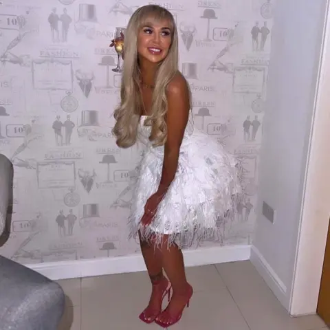 Janet Taylor Louise - a young woman with long straight blond hair - posing at home in a white party dress and pink high-heeled shoes. She is holding a glass of white wine and standing next to a wall with grey patterned wallpaper.