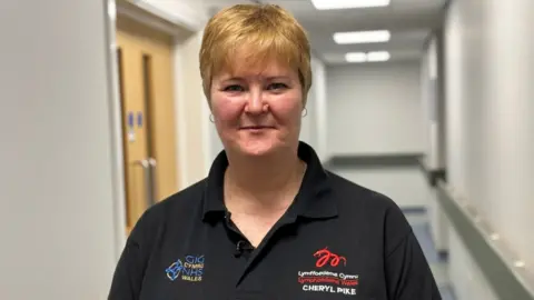 A white woman with short blonde hair and black NHS branded polo top looking at the camera