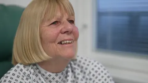 Margaret Georgiou is chatting to a BBC reporter and is dressed in a surgical patient gown