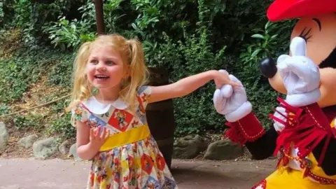 Anna Cieslik Beatrice, with blonde pigtails, smiling and holding out her hand to Mickey Mouse at Disneyland. She is wearing a patchwork dress featuring Toy Story characters.