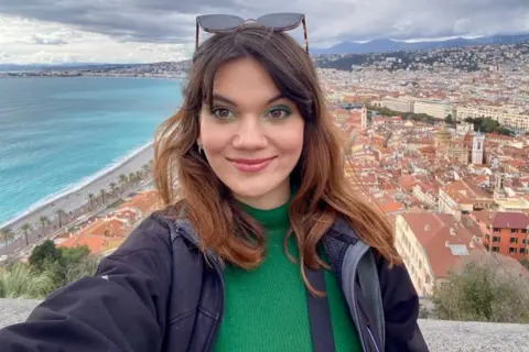 Bianca Ionici A woman is smiling for a selfie from a tower. These is a city, mountains and the sea in the background