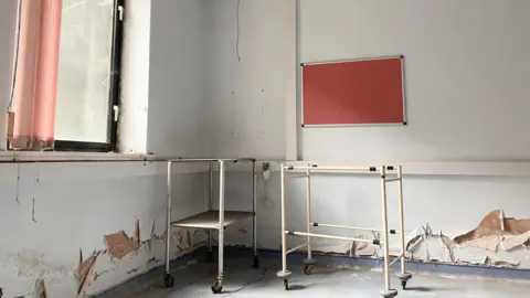 An abandoned clinic is pictured with a red noticeboard and peeling wallpaper visible