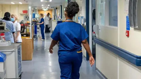 PA Media A nurse in blue scrubs walks through a busy hospital corridor