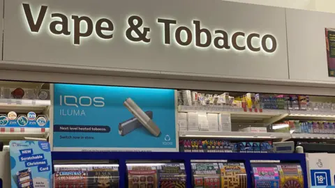 BBC Vapes and tobacco kiosk in a Sainsbury's store in London. Sign reads 'vape and tobacco'. Below is a blue sign advertising iQos Iluma, "Next level heated tobacco", with the words "Switch now in store". 