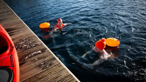 Emma Lynch/BBC James treads water at the half-way point of his swim. Only his head is above water and he is wearing an orange bobble hat. A swimming buoy is attached to him. There is another swimmer in the water next to him.