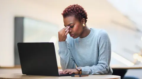 Getty Images Stock image of a woman sitting at a laptop looking down with her head in her hand. She has short hair and is wearing a blue-grey jumper.