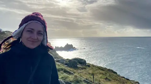 Emily Jones Emily Jones wearing a woollen hat stands on a clifftop path with the ocean behind her