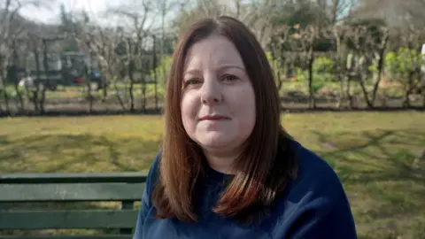 Headshot of Lucy. She has long, dark, straight hair and is wearing a navy blue sweatshirt. She is photographed outside, on a green bench, with shrubs behind her.