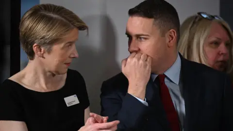 PA Media Amanda Pritchard, wearing a black top with a name tag on, stands speaking to West Streeting, who is wearing a suit. Pritchard is gesturing with her hands and Streeting is covering his mouth with his hand