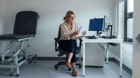 Getty Images A woman at a computer reads a document on paper. She is sitting in an officer chair and there is a medical bed behind her.