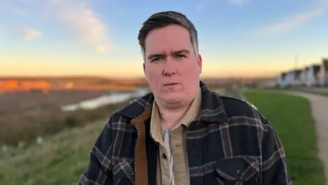 Kate Bradbrook/BBC A man in a beige shirt, and navy tartan coat, stands on a strip of grass, looking straight at the camera.