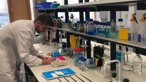 University of Northampton A man in a white lab coat and glasses writes on a table with lots of lab equipment on a large white table in front of him. 