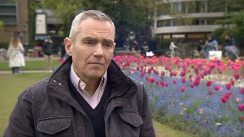 Ignacio Labayen De Inza, a man with grey hair wearing a dark coloured coat over a dark coloured V-neck jumper and pink shirt, stood in front of pink and blue flowers